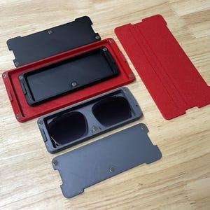 May include: A set of sunglasses with a gray frame and dark lenses, inside a gray case. The case is open, revealing the glasses. A red case and a black case are also visible, along with a red cover.