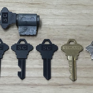 Può includere: Un assortimento di chiavi e un cilindro di serratura sono esposti su una superficie di legno. Le chiavi includono diverse chiavi nere con "SC", una chiave in ottone con "SCHLAGE" e una chiave d'argento con "HILLMAN".