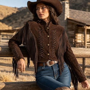 May include: A dark brown suede jacket featuring long fringe detailing along the sleeves and chest, accented with vertical lace-up cord patterns and round metallic buttons. The jacket is paired with blue denim jeans and a brown leather belt.