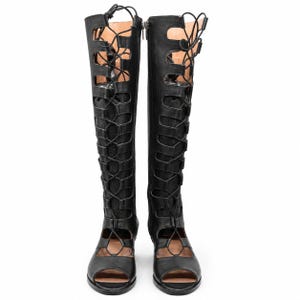 May include: A pair of black knee-high gladiator sandals featuring an open-toe design, a tall shaft with cut-out panels, and a full-length lace-up closure system that runs from the top of the shaft down to the foot area with thin black laces.