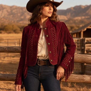 May include: A deep burgundy suede jacket featuring long fringe detailing on the sleeves and shoulders, a high collar, and decorative lace-up accents on the front and cuffs. The jacket is fastened with metallic buttons and worn over a light shirt.