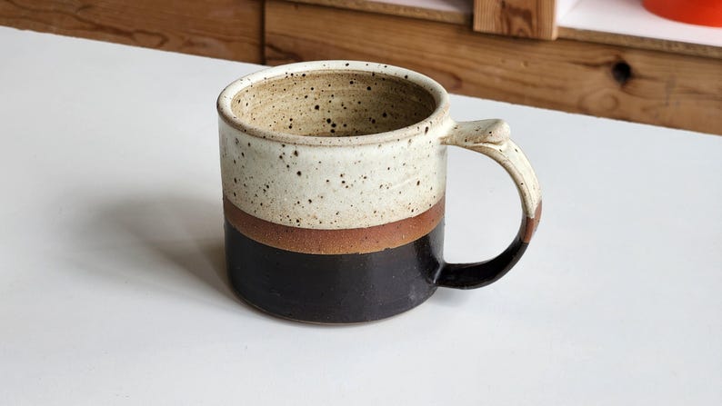以下が含まれることがあります： 手作りの陶器マグカップ。上部は斑点のあるクリーム色、中央に茶色の帯、下部は濃い茶色です。ハンドルはクリーム色で、濃い茶色のアクセントが付いています。コーヒーや紅茶に最適です。