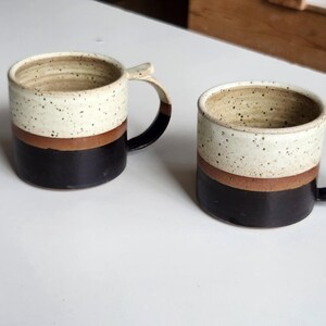 以下が含まれることがあります： 白の斑点模様のトップ、茶色の帯、黒色のベースのセラミックマグカップが2つ。各マグカップには湾曲したハンドルが付いています。マグカップは白い表面にあります。