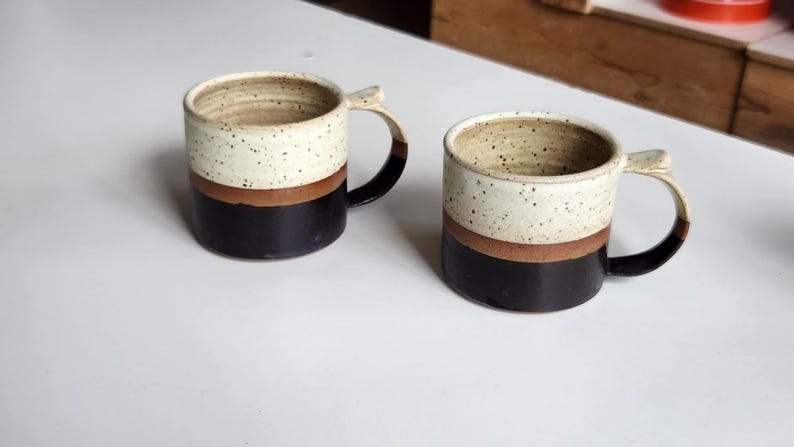 以下が含まれることがあります： 白の斑点模様のトップ、茶色の帯、黒色のベースのセラミックマグカップが2つ。各マグカップには湾曲したハンドルが付いています。マグカップは白い表面にあります。