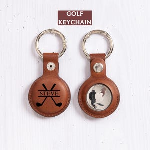 May include: Two brown leather golf keychains with silver key rings. One keychain features crossed golf clubs and the name "STEVE". The other keychain has a golf-themed design. The text "GOLF KEYCHAIN" is at the top.