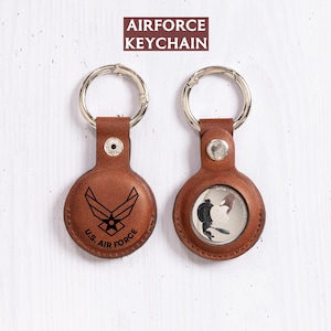 May include: Two brown leather Air Force keychains with silver key rings. One keychain features the Air Force logo and text, the other has a black and white image. The text "AIRFORCE KEYCHAIN" is at the top.
