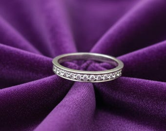 May include: A silver ring with a band of small, clear gemstones. The ring is set against a backdrop of deep purple velvet fabric. The gemstones are evenly spaced around the band, creating a sparkling effect. The ring is a simple, elegant design.