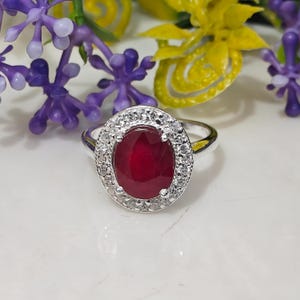 Peut inclure: Une bague en argent ornée d'un grand rubis ovale au centre, entouré d'un halo de petits diamants transparents. La bague est posée sur une surface blanche avec des accents floraux violets et jaunes.