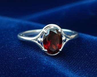 May include: A silver ring with an oval, deep red gemstone. The ring's setting has an ornate design with small decorative elements on either side of the gemstone. The ring is set against a dark blue background.