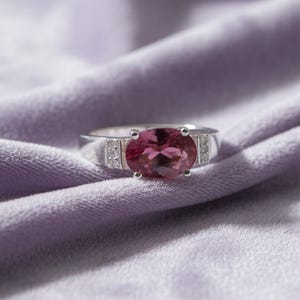 May include: A silver ring with a large, oval-cut, pink gemstone as the centerpiece. The ring has small, clear accent stones on either side of the main stone, set against a soft, purple fabric background.