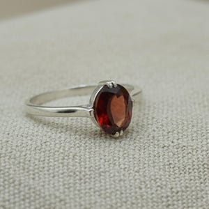 May include: A silver ring featuring an oval-cut, deep red gemstone. The gemstone is set in a four-prong setting, and the band is a simple, polished silver. The ring is displayed on a neutral-toned surface.