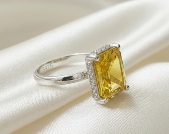 May include: A silver ring with a large, rectangular yellow gemstone. The gemstone is surrounded by smaller, clear stones. The ring band is thin and has the marking "S925". The ring is set against a white, silky background.