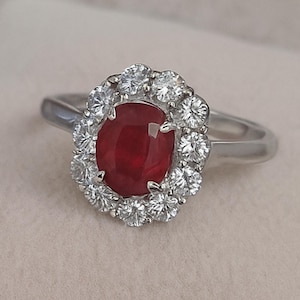 Peut inclure: Une bague en argent ornée d'un rubis ovale central, entouré d'un halo de petits diamants ronds et transparents. Le rubis est d'un rouge profond, et la bague a un anneau en argent poli.