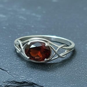 May include: A silver ring with an oval, dark red gemstone. The ring has a decorative, interwoven design around the gemstone. The band is a simple, polished silver. The gemstone is approximately 0.5 inches in length.