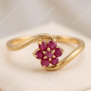 Peut inclure: Une bague en or ornée d'un motif floral composé de sept rubis ronds rose foncé. L'anneau présente une finition polie et un design croisé qui entoure la composition centrale de pierres précieuses, formant une boucle continue.