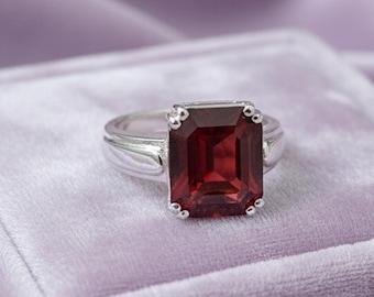 Könnte beinhalten: Ein silberner Ring mit einem großen, rechteckigen, tiefroten Edelstein. Der Edelstein ist der Mittelpunkt, eingebettet in ein klassisches, elegantes Design. Der Ring wird auf einer weichen, violetten Samtoberfläche präsentiert.