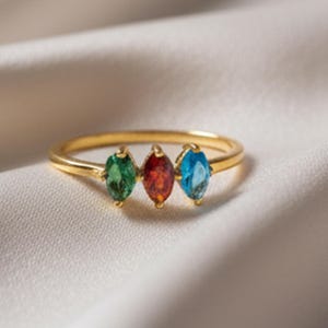 Pode incluir: Um anel dourado com três pedras preciosas em forma de marquesa nas cores verde, vermelho e azul. As pedras estão dispostas em fila numa fina banda dourada. O anel é exibido numa superfície branca texturizada.