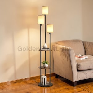 May include: A black floor lamp with three lampshades and built-in shelves. The lamp has a round base and a slender, vertical design. The shelves hold a small plant, books, and decorative items. The lampshades are beige, and the text "Golden WL" is visible.