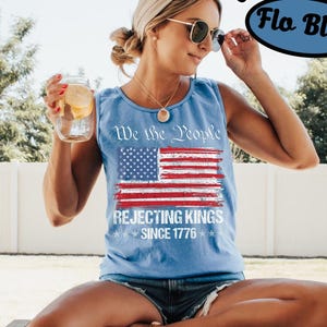 May include: A light blue sleeveless tank top featuring a distressed American flag graphic with the text We the People REJECTING KINGS SINCE 1776 printed in white. The shirt has a crew neckline and a relaxed fit, showcasing a patriotic design.