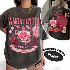 以下が含まれることがあります： ピンク色の「AMORTENTIA」の文字が入った濃いグレーのコンフォートカラーTシャツ。前面には、王冠、バナー、その他のグラフィックが描かれたハート型のデザインが施されています。背中には、テキストとキャンドルが付いた同様のデザインがあります。