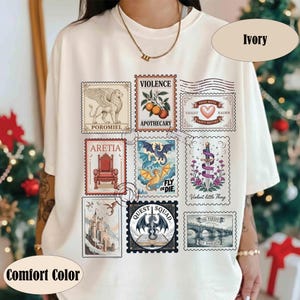K&ouml;nnte beinhalten: Elfenbeinfarbenes T-Shirt mit einer Reihe von Briefmarken-Designs zum Thema Fantasy. Die Briefmarken zeigen einen Greif, einen Drachen, ein Schloss und Texte wie "Violence Apothecary" und "Quest Squad". Das T-Shirt ist mit "Comfort Color" gekennzeichnet.