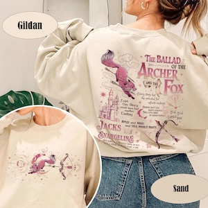 May include: Sand-colored Gildan sweatshirt featuring a graphic design with the text "The Ballad of the Archer and the Fox." The design includes a pink fox with arrows, a castle, and other illustrations.