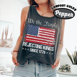 May include: A charcoal gray sleeveless tank top featuring a distressed American flag graphic. The text reads We the People, REJECTING KINGS, and SINCE 1776. The shirt is labeled as comfort color pepper, showcasing a vintage-style print design.