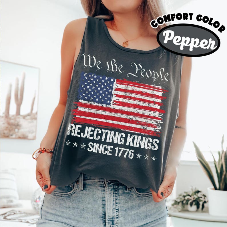 May include: A charcoal gray sleeveless tank top featuring a distressed American flag graphic. The text reads We the People, REJECTING KINGS, and SINCE 1776. The shirt is labeled as comfort color pepper, showcasing a vintage-style print design.