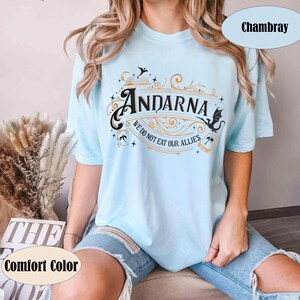 May include: Light blue t-shirt with the word "Andarna" in a decorative font, along with the phrase "We do not eat our allies." The shirt features a fantasy-themed design with dragons and stars. The shirt is labeled "Comfort Color" and "Chambray".