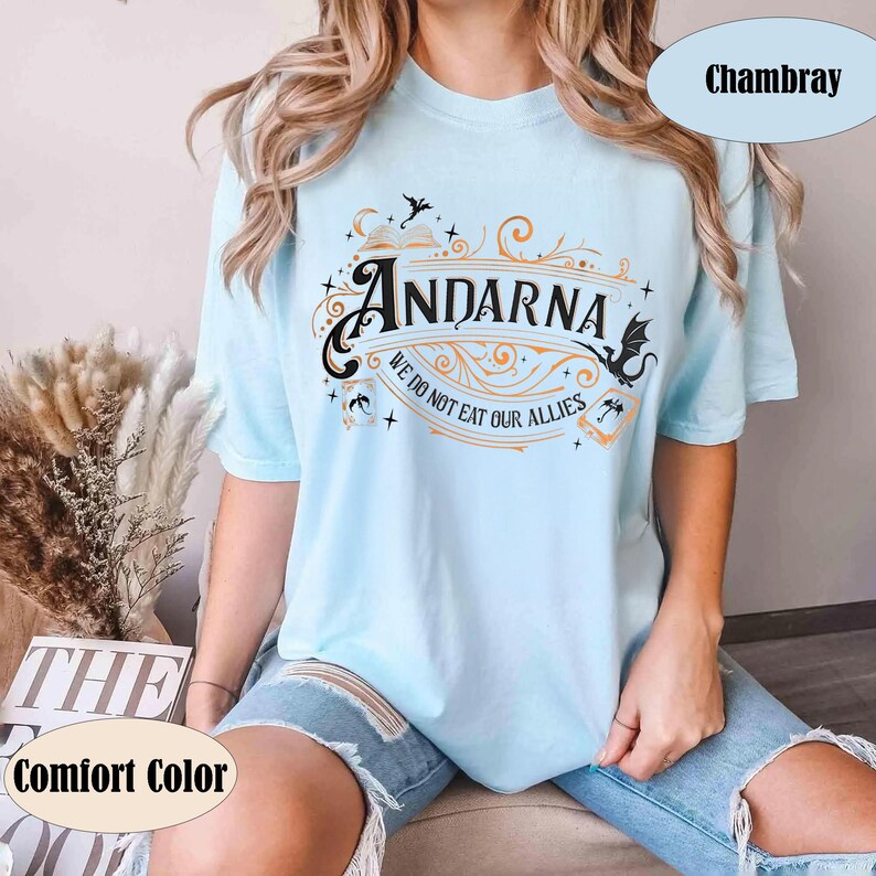 May include: Light blue t-shirt with the word "Andarna" in a decorative font, along with the phrase "We do not eat our allies." The shirt features a fantasy-themed design with dragons and stars. The shirt is labeled "Comfort Color" and "Chambray".