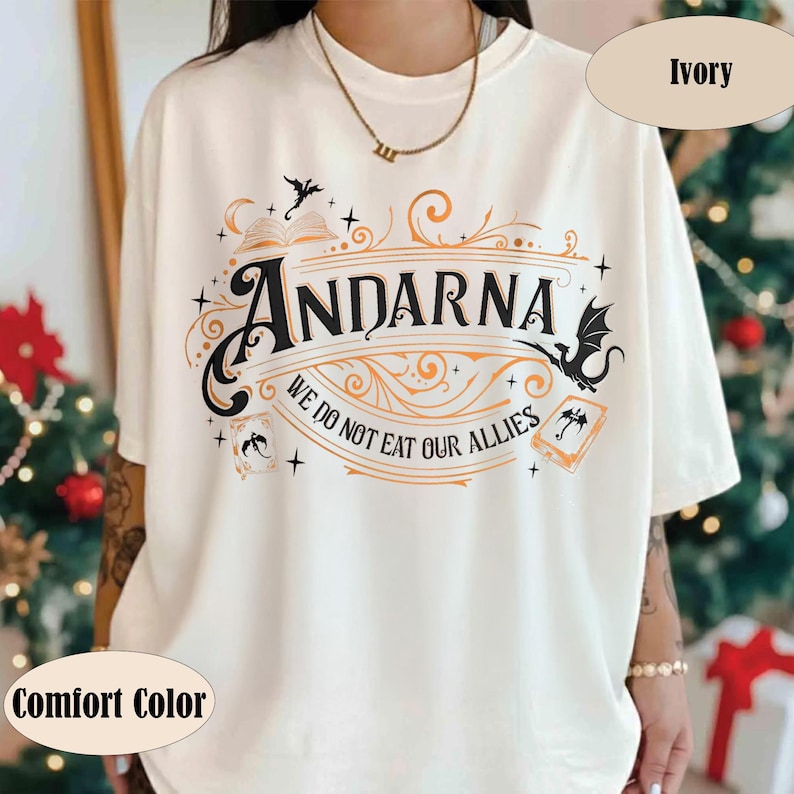 May include: Ivory-colored t-shirt with the word "ANDARNA" in a decorative font, accompanied by the phrase "WE DO NOT EAT OUR ALLIES." The design includes illustrations of dragons, books, and stars. The shirt is labeled "Comfort Color."