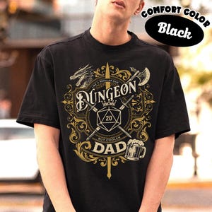 May include: Black t-shirt featuring a gold and white graphic. The design includes the word "Dungeon", a dragon, a crown, a 20-sided die, crossed swords, and the words "Like a regular dad but cooler dad".