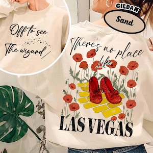 May include: Sand-colored sweatshirt with the text "Off to see The wizard" on the front. The back features red shoes, poppies, and the text "There's no place like Las Vegas."
