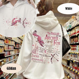 May include: White hoodie with a whimsical design featuring a pink fox with a bow and arrow, and the text "The Ballad of the Archer and the Fox." The design includes a castle, stars, and other illustrations. The hoodie is made by Gildan.