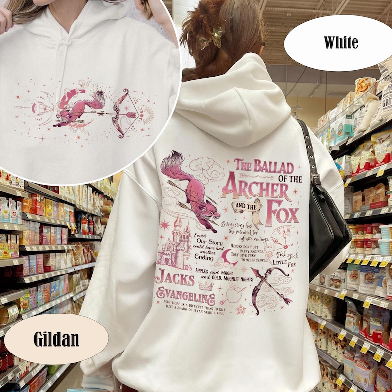 May include: White hoodie with a whimsical design featuring a pink fox with a bow and arrow, and the text "The Ballad of the Archer and the Fox." The design includes a castle, stars, and other illustrations. The hoodie is made by Gildan.