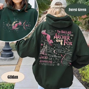 May include: Dark green hoodie with a pink fox and archer graphic, and the text "The Ballad of the Archer and the Fox." The hoodie has a hood with a drawstring and a front pocket. The brand name "Gildan" is visible.