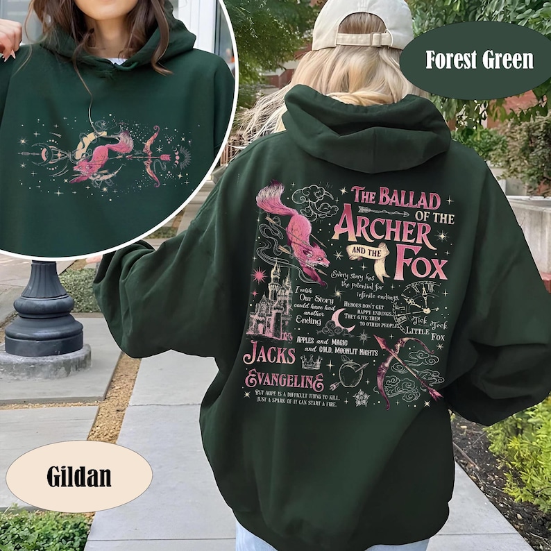 May include: Forest green hoodie with a graphic featuring a pink fox and archer design, with text that reads "The Ballad of the Archer and the Fox." The hoodie has a drawstring hood and a front pocket. The brand name "Gildan" is visible.