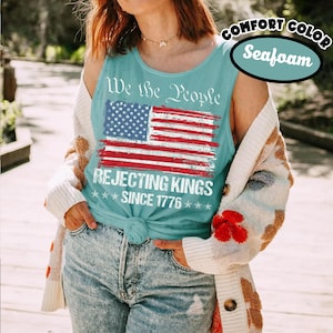 May include: A seafoam green tank top featuring a distressed American flag graphic with the text We the People, REJECTING KINGS, and SINCE 1776 printed in white. The tank top is worn with a cream-colored cardigan and blue denim jeans.