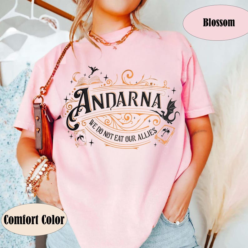 May include: A light pink t-shirt with the word "Andarna" in a decorative font, along with the phrase "We do not eat our allies." The shirt has a graphic design with bats and other fantasy elements. The shirt is labeled "Comfort Color" and "Blossom".