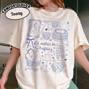 Op de afbeelding: Ivoor Comfort Color T-shirt met een blauw grafisch ontwerp. Het ontwerp bevat illustraties van kopjes, borden, een winkelwagentje en de tekst "I'd rather be Thrifting". Het shirt heeft een relaxte pasvorm.