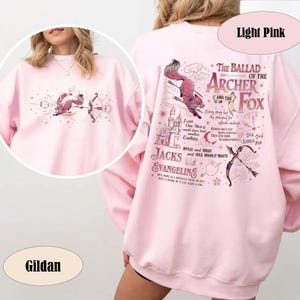 May include: Light pink Gildan sweatshirt featuring a whimsical design. The front displays a fox with a bow and arrow, while the back showcases text and illustrations of a fox and archer. The text includes "The Ballad of the Archer and the Fox."