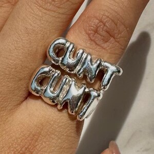 以下が含まれることがあります： 「CUNT」の文字が2段に重ねられたシルバーリング。指にはめられており、文字は太字のバブルスタイルフォントです。リングは光沢のある金属製です。