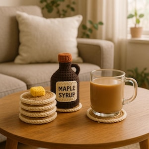 Op de afbeelding: Een stapel gehaakte pannenkoeken met een gele klont boter, een bruine ahornsiroopfles met een label "MAPLE SYRUP" en een glazen mok koffie op een houten tafel. De items zijn handgemaakt.
