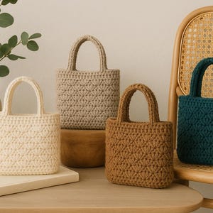 Könnte beinhalten: Vier kleine, handgefertigte Häkeltaschen in Creme, Beige, Braun und Blaugrün. Jede Tasche hat eine strukturierte Form mit einem Griff und einem strukturierten, blütenartigen Muster. Die Taschen werden auf einer Holzoberfläche mit einer Vase mit Grün und einem Korbstuhl ausgestellt.