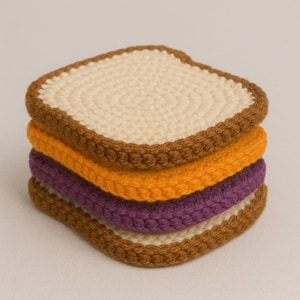 May include: A stack of four crocheted coasters resembling a sandwich. The top and bottom layers are off-white with brown edges, while the middle layers are orange and purple. The coasters are square with rounded corners.