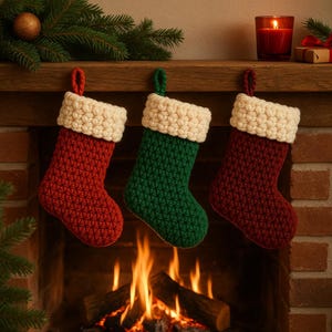 Puede incluir: Tres calcetines navideños en rojo, verde y burdeos, cada uno con un puño y lazo color crema para colgar. Los calcetines se exhiben frente a una chimenea con una vela encendida y vegetación.