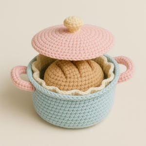 Puede incluir: Una olla de juguete de ganchillo con tapa, asas y una hogaza de pan en el interior. La olla es azul claro con asas rosas, una tapa rosa y un interior beige. El pan es marrón claro. La tapa tiene un pomo amarillo.