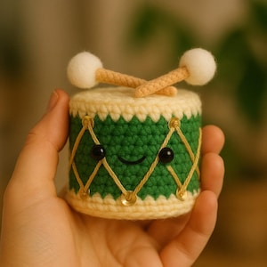 Puede incluir: Un tambor de ganchillo hecho a mano con una cara sonriente, ojos de cuentas negras y un diseño cosido en amarillo. El tambor es verde con la parte superior e inferior de color crema. Dos baquetas marrones con puntas blancas descansan encima.