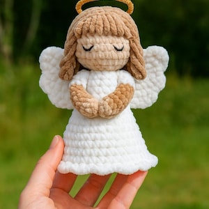 May include: A handmade crochet angel doll with a white dress, brown hair, and a gold halo. The angel has closed eyes and folded hands, and white wings. The doll is held in a person's hand against a blurred green background.