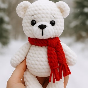 May include: A handmade white crocheted bear with black eyes and a black nose. The bear is wearing a red scarf. The bear is being held in a person's hand. The background is a snowy forest.
